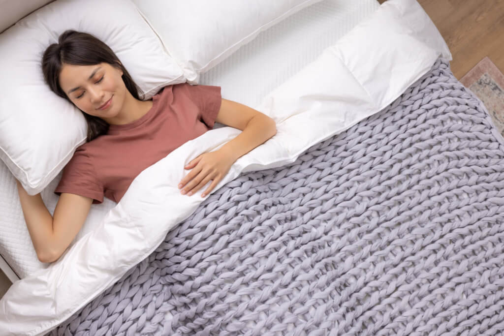 A woman sleeping happily in bed underneath a light grey braided weighted blanket